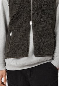 Veste en polaire marron foncé avec fermeture éclair sur le devant, deux poches latérales et une surface texturée, portée sur un sweat-shirt gris et un pantalon en velours côtelé noir.
