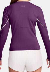 Žena v dlouhém sportovním topu fialové barvy s bílými detaily a bílých skládacích šortkách, zobrazená zezadu.