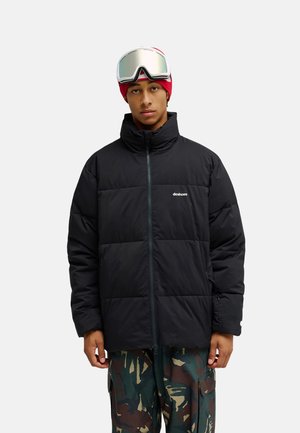 Jonge persoon die een zwarte donsjacket, rode muts, reflecterende skibril en camouflerende broek draagt, staand tegen een effen achtergrond.