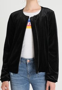 Svart sammet bomberjacka med rund halsringning, dragkedjeslutning och elastiska muddar. Bärs över en vit t-shirt och tillsammans med blå jeans.