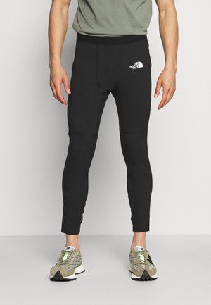 Schwarze sportliche Leggings mit figurbetontem Design, glatter Textur und einem Logo am oberen Oberschenkel. Kombiniert mit hellen Laufschuhen.