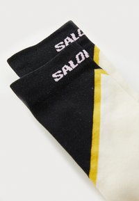 Sorte, cremefarvede og gule sokker med et geometrisk design, der har et rosa "SALON" logo på kanten. Materialet er en bomuldsblanding med struktureret overflade.