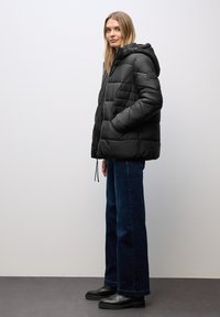 Giacca a vento nera con cappuccio, caratterizzata da un design trapuntato e cordini elastici. Indossata sopra jeans a gamba larga blu e stivali neri.