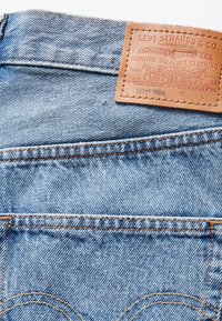 Jeans de mezclilla azul claro con una superficie texturizada. Parche de logo de cuero marrón cosido en la cinturilla. Detalles de costura naranja visibles.