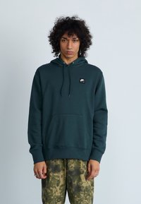 Sweatshirt verde escuro com um bolso frontal e cordões. Apresenta um pequeno logótipo preto da Nike Air. Usado com calças verdes estampadas.