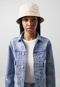 Giacca in denim con ricamo geometrico multicolore, bottoni, due tasche laterali; abbinata a un cappello a secchiello beige in un tessuto liscio e leggero.