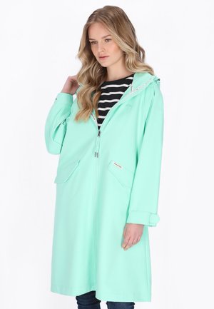 Mujer con cabello largo y rubio, vestida con un impermeable verde menta con capucha sobre una camiseta de rayas blancas y negras, posando contra un fondo blanco.