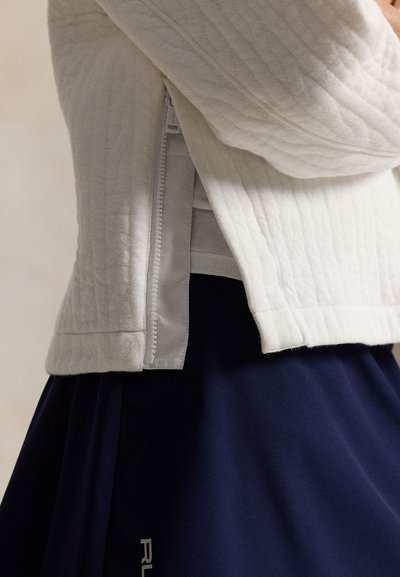 Λευκό μπουφάν με καπιτονέ ραφή και λεπτομέρεια πλαϊνού φερμουάρ, φορεμένο πάνω από γκρι μπλουζάκι και συνδυασμένο με φούστα σε σκούρο μπλε χρώμα.