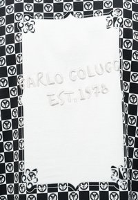 Λευκό ορθογώνιο έμπλαστρο με μαύρες διακοσμητικές γωνίες και επαναλαμβανόμενο μοτίβο με σύμβολο ειρήνης στο φόντο, κεντημένο με τη φράση «Carlo Colucci Est. 1976.»