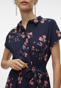 Kvinna i en marinblå klänning med rosa blommönster, knuten i midjan, och långa pärlörhängen, justerar midjebandet.