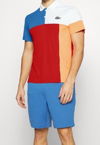 Koszulka polo w blokach kolorów: niebieski, czerwony, pomarańczowy i biały. Do tego jasnoniebieskie spodenki z teksturowanym wzorem.