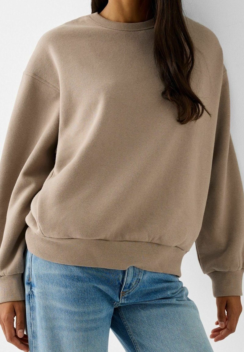 Femme portant un sweat-shirt beige à manches longues et un jean en denim bleu clair, debout devant un fond clair uni.