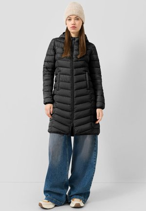 Femme portant une longue doudoune noire, un jean bleu à jambes larges, un bonnet beige en tricot et des baskets blanches, debout sur un fond gris uni.