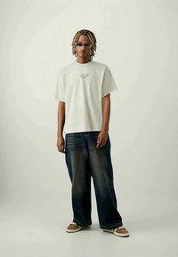 Camiseta de algodón blanca de manga corta, con un gráfico impreso en el pecho, combinada con jeans de denim azul oscuro de corte holgado y zapatillas.
