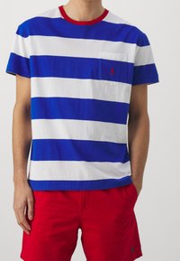 Bomullströja med blå och vita horisontella ränder, röd ribbad krage, ficka på vänster bröst, matchad med röda shorts.