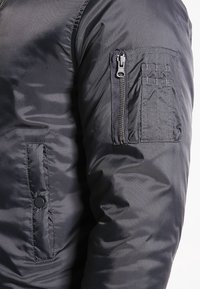 Mörkgrå jacka med ärm som har en zippered fack och sydda fack, visar tygtextur och sömnadsdetaljer.