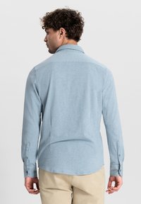 Camicia azzurro chiaro a maniche lunghe con colletto button-down, caratterizzata da una texture morbida e polsini a due bottoni, indossata sopra pantaloni khaki.