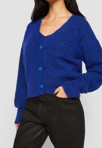 Blå virkade cardigan med V-ringning, texturerat tyg, och fyra blå knappar fram. Matchad med svarta byxor.