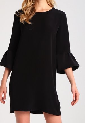 Femme portant une robe noire mi-longue aux genoux avec des manches cloche trois-quarts et un col rond sur un fond clair uni.