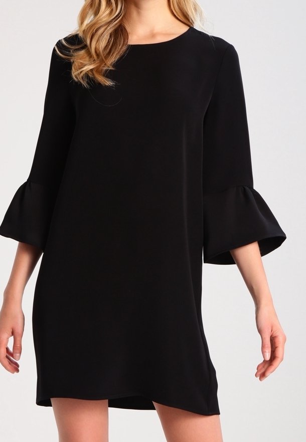 Femme portant une robe noire mi-longue aux genoux avec des manches cloche trois-quarts et un col rond sur un fond clair uni.