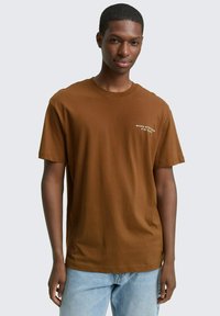 Bruine katoenen t-shirt met korte mouwen, ronde hals, en witte tekstaccenten op de linkerborst. Gedragen met lichtblauwe spijkerbroek.