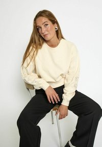 Giovane donna con i lunghi capelli biondi, che indossa un sudadera crema con dettaglio in pizzo sulle maniche, pantaloni neri e orecchini d'argento, seduta su una sedia di metallo.
