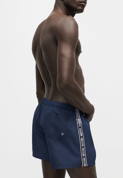 Άνδρας χωρίς μπλούζα που φοράει σκούρα μπλε μαγιό με λευκή λωρίδα λογότυπου Moschino στα πλάγια, στέκεται με τα χέρια στους γοφούς μπροστά σε λευκό φόντο.