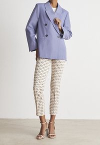 Lavendel dobbeltknappet blazer og mønstrede cremede bukser med geometriske accenter. Modellen har sandfarvede stiletter på. Glatte teksturer gennemgående.