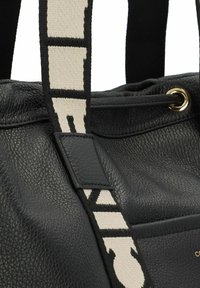 Sac à main en cuir noir avec une surface texturée, doté d'une large bandoulière crème avec des lettres noires, et un accent de ferrure ton or.