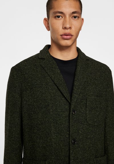 Grönullig blazer med en texturerad yta, snedskurna slag, två knappar och en bröstficka. Buren över en svart T-shirt.