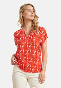 Rode blouse met een V-hals en korte mouwen, met een draaiend abstract patroon in wit en roze op een gestructureerde stof.