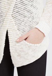 Gilet blanc en tricot avec un tissage texturé, présentant un devant drapé et deux poches latérales. Manches arrondies et design léger.