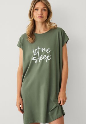 Blondynka w luźnej zielonej sukience z krótkim rękawem z białym napisem "let me sleep", stojąca na jednolitym jasnym tle.