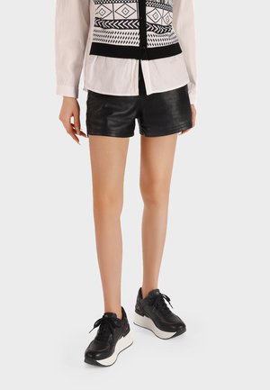 Donna che indossa pantaloni corti in pelle nera, gilet a motivi bianco e nero, camicia bianca e sneakers platform nere mentre sta in piedi.