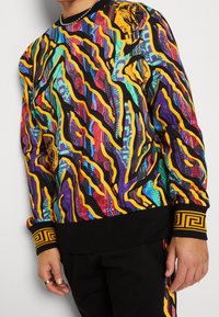 Flerfärgad sweatshirt med djärva abstrakta mönster i lila, blått, gult och svart. Har ett svart band med en gyllene grekisk nyckeldesign vid ärmsluten.
