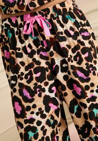 Pantalons de pyjama à imprimé léopard en tissu léger avec un cordon de serrage rose. Présente de grandes taches noires, brunes et colorées sur un fond beige.