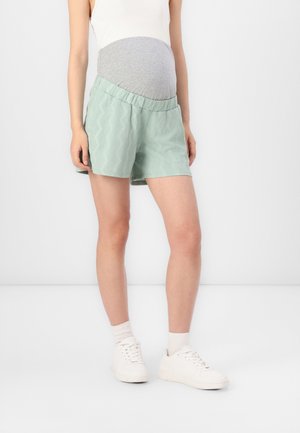 Persona embarazada con pantalones cortos de maternidad verde claro con un panel gris, camiseta sin mangas blanca, calcetines blancos y zapatillas blancas, de pie en interiores.