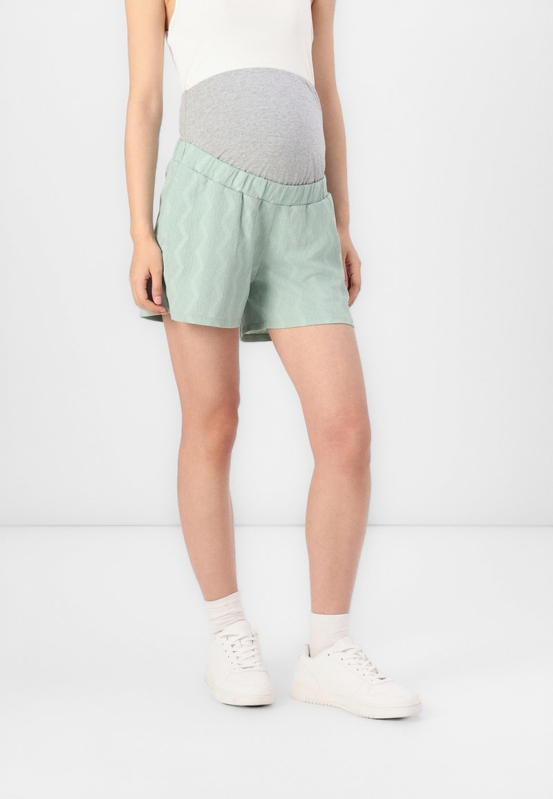 Personne enceinte portant un short de maternité vert clair avec un panneau gris, un débardeur blanc, des chaussettes blanches et des baskets blanches, debout à l'intérieur.