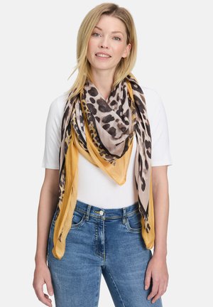 Blonde vrouw die een wit shirt met korte mouwen, een blauwe spijkerbroek en een grote geelbruine sjaal met luipaardprint om haar nek draagt.