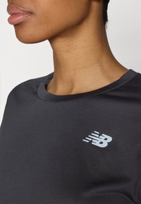 Läheltä kuvattu henkilö, jonka kaula ja alaleuka näkyvät, yllään tummanharmaa paita, jossa on valkoinen New Balance -logo rinnassa.