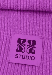 Tessuto lavorato a maglia viola con coste verticali. Presenta un'etichetta in viola abbinato, che mostra un logo e la parola "STUDIO" in viola scuro.