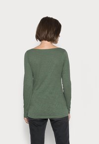 Groene blouse met lange mouwen, gemaakt van een gestructureerde, semi-doorzichtige stof en een wijde halslijn, vanaf de achterkant weergegeven. Gecombineerd met donkere broeken.