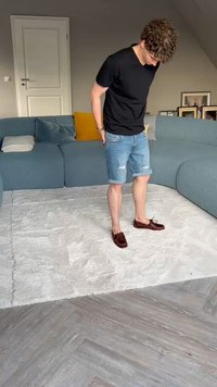 T-shirt preto, calções de ganga com bordas desfiadas e mocassins castanhos. Tapete de cor clara e sofá seccional estofado ao fundo.