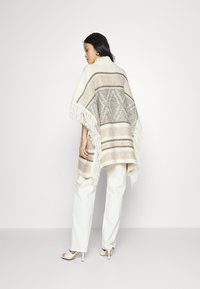Poncho de malha com padrões geométricos em bege e creme, apresentando franjas ao longo da borda. Combinado com calças brancas e sandálias de salto alto.