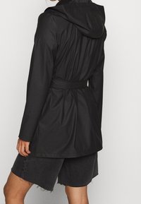 Veste imperméable noire avec capuche, taille ceinturée et manches longues. Caractérisée par une texture lisse et un design resserré à l'arrière.