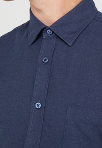 Närbild av en marinblå skjorta med krage, tre synliga knappar och en bröstficka på högra sidan.