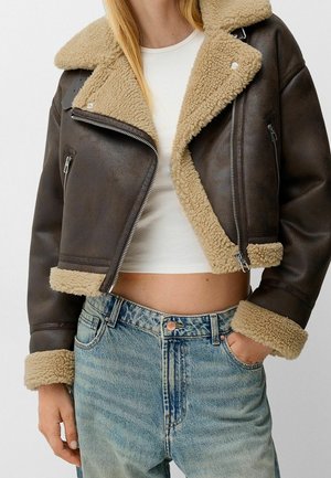 Kobieta w brązowej, krótkej kurtce ze skóry z kożuszkiem, białym, krótkim topie i jasnoniebieskich, wysokich dżinsach, z ręką w kieszeni.