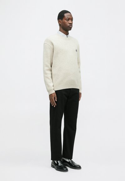 Bežs vilnas džemperis ar ribotām piedurknēm un apakšmalu. Melni bikses, ādas kurpes. Models stāv uz vienkārša balta fona.