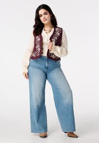 Bordeauxrode geborduurde vest over een crèmekleurige blouse, gecombineerd met lichtblauwe wijde jeans en beige hakken. Soepele texturen en bloemmotieven.