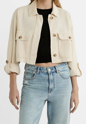 Vrouw draagt een korte crèmekleurige jas met opgerolde mouwen, een zwarte korte top en lichtblauwe hooggetailleerde jeans.